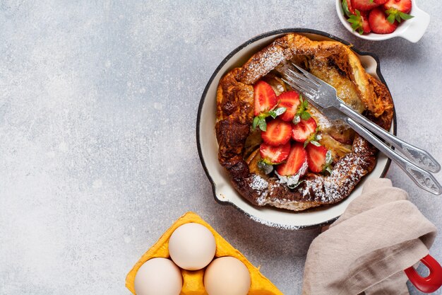 Nederlandse babypannenkoek met verse aardbeibes en bestrooid met poedersuikerpoeder in rode pan op witte keukenachtergrond. Bovenaanzicht.