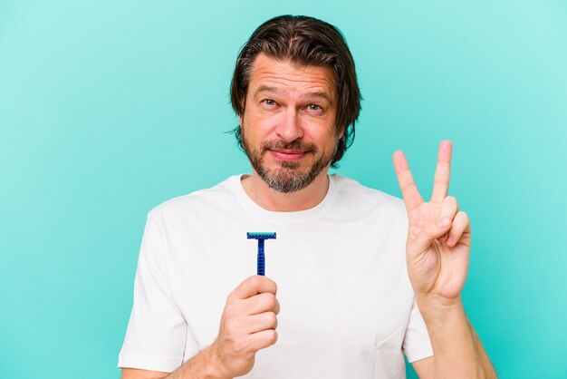 Foto nederlandse man van middelbare leeftijd die een scheermesje houdt dat op blauwe muur wordt geïsoleerd die nummer twee met vingers toont.