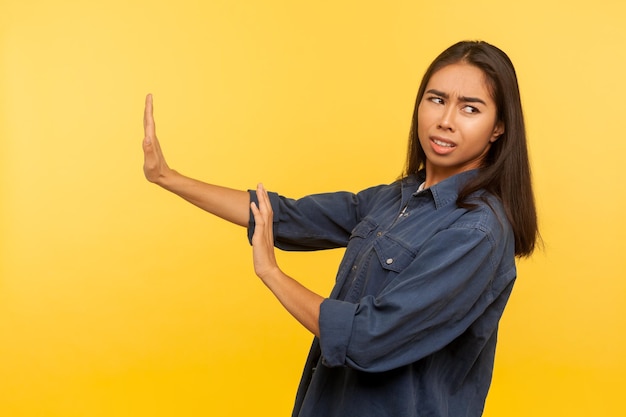 Nee I39m bang zijaanzicht van bang meisje in denim shirt staande met opgeheven handen stop gebaar in paniek op zoek bang voor gevaar fobie indoor studio opname geïsoleerd op gele achtergrond