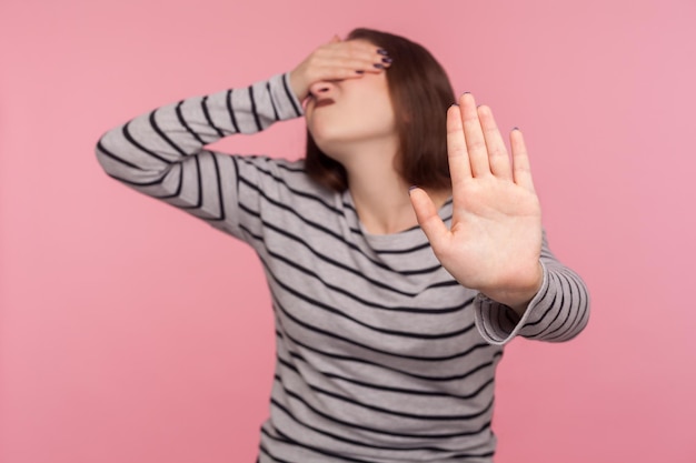 Nee, ik wil hier niet naar kijken. Portret van een vrouw in een gestreept sweatshirt die de ogen bedekt en zich met afschuw omdraait van iets beschamends, met een stopgebaar. studio-opname geïsoleerd op roze achtergrond