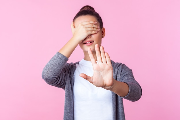 Nee, ik wil niet kijken. Portret van een brunette tienermeisje met een knotkapsel in vrijetijdskleding die de ogen bedekt en een stopgebaar toont, bang of geschokt door beschamende inhoud. studio-opname binnenshuis