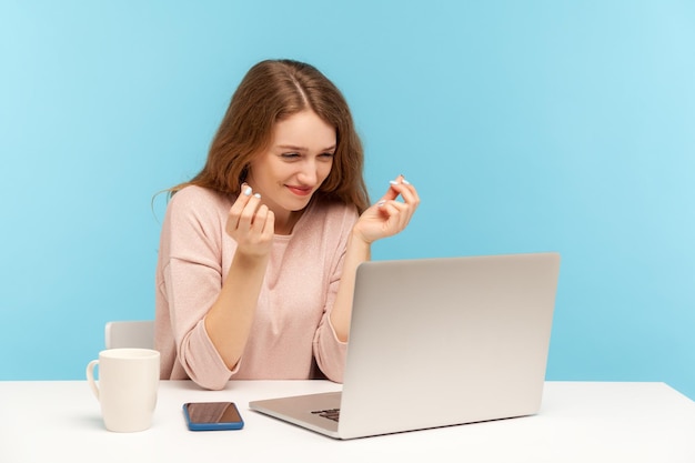 Need higher salary. cheerful young woman employee showing money gesture to laptop screen, discussing payment on video call, planning business income. indoor studio shot isolated on blue background