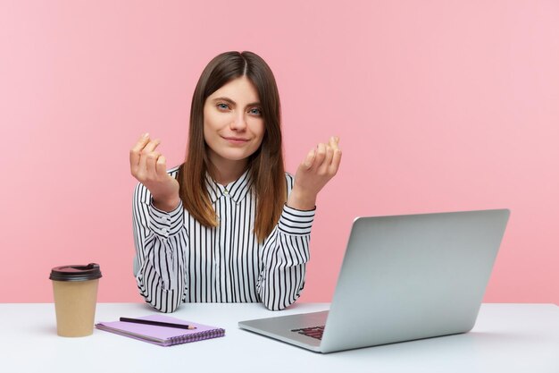 Need higher salary Positive woman office worker sitting at workplace with laptop and showing money gesture asking payment planning business income Indoor studio shot isolated on pink background