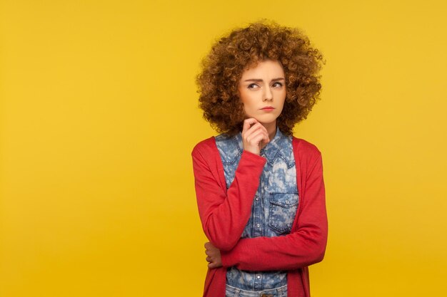 Need to think. Portrait of pensive woman with fluffy curly hair in casual outfit holding chin and pondering idea, planning strategy, musing and solving difficult question in mind. studio shot isolated