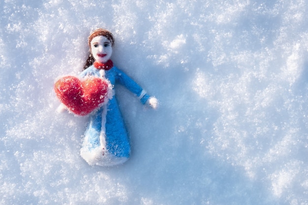 Foto bambola di lana infeltrita ad ago sdraiata sulla neve con un cuore rosso
