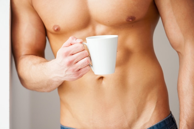 Neem even de tijd om te ontspannen! Close-up van een jonge gespierde man met een perfecte torso die een kopje koffie vasthoudt terwijl hij tegen de muur leunt