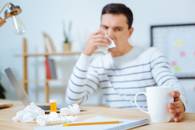 Negative thoughts. Sick young man looking at his cup and holding napkin near nose while sitting at his workplace