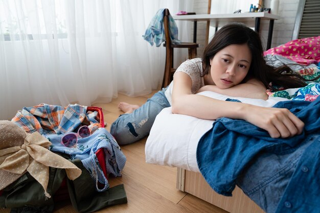 negative young asian woman in trouble relying on bed near suitcase on wooden floor. worried tired lady upset packing luggage in bedroom. unhappy female preparing for vacation holidays at home.