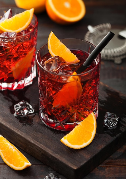 Negroni cocktail in crystal glasses with orange slice and fresh raw oranges on chopping board with strainer on wooden background Top view