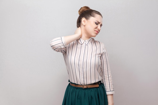 Nek pijn. Portret van ongelukkige mooie jonge vrouw in gestreept shirt en groene rok en verzameld verbodskapsel, staande en haar pijnlijke nek vasthoudt. studio opname, geïsoleerd op een grijze achtergrond.