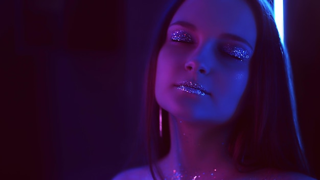 Портрет в неоновом свете. Креативный модный макияж. Женщина с блестящими губами закрыла глаза сине-фиолетовым свечением.