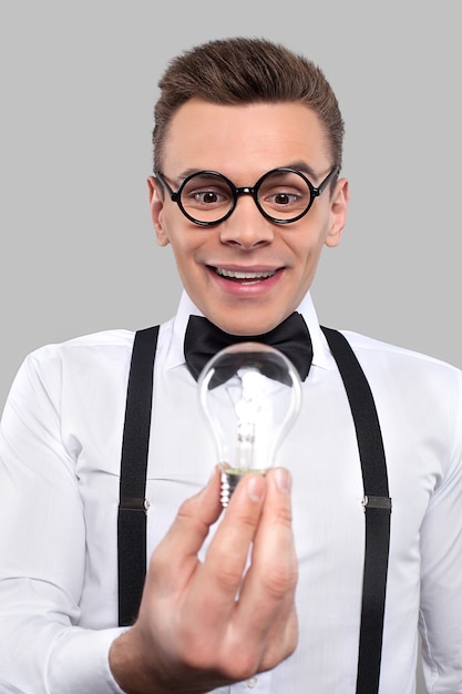 Nerd with a light bulb. Surprised young man in bow tie and suspenders holding a light bulb and looking at it while standing against grey background