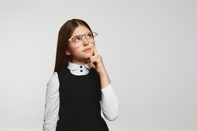 Foto ragazza nerd con gli occhiali e l'uniforme scolastica che sfrega il mento e distoglie lo sguardo mentre pensa