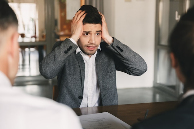 Nerveus gespannen man 30s zorgen maken en zijn hoofd grijpen tijdens sollicitatiegesprek op kantoor, met collectief van specialisten - bedrijfs-, carrière- en wervingsconcept