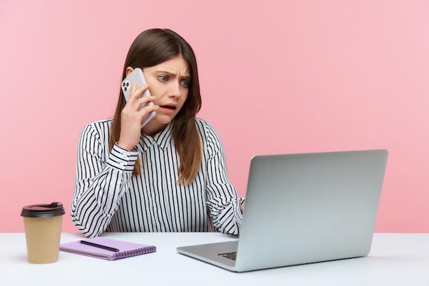 Nerveus ongelukkige vrouw kantoormedewerker praten telefoon met client zit op werkplek manager bestelling maken op laptop boeking tickets Indoor studio opname geïsoleerd op roze achtergrond