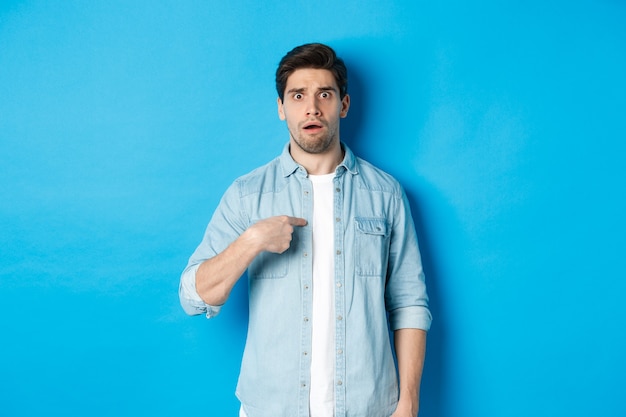 Foto nerveuze man wijzend naar zichzelf en kijkt verward, staande in vrijetijdskleding over blauwe muur