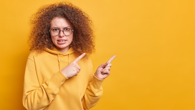 Nervous displeased curly haired woman frowns face says its unfair points at upper right corner with dislike expression complains about something bad wears glasses sweatshirt isolated on yellow wall