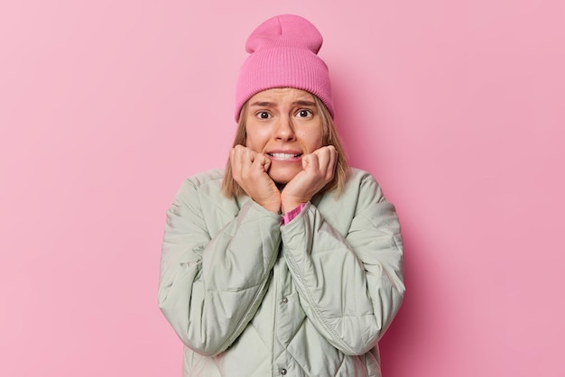 Nervous embarrassed European woman keeps hands under chin trembles from fear afraids of speaking stands insecure wears hat and jacket isolated over pink background. Worried girl feels anxious