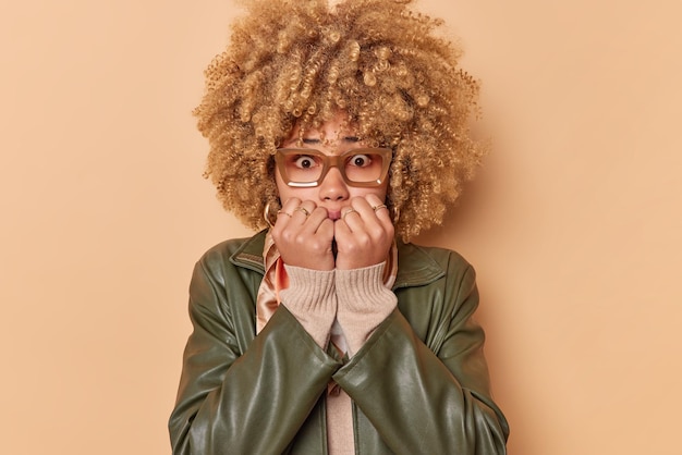Nervous embarrassed woman bites finger nails feels scared reacts on something looks anxious wears spectacles and leather jacket isolated over beige studio background has anxiety or being troubled
