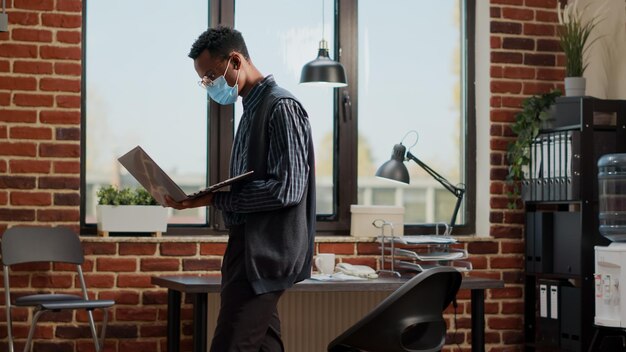 Nervous employee waiting to receive important news at work, pacing around office with laptop in hand. Impatient consultant expecting news about executive career, wearing face mask.