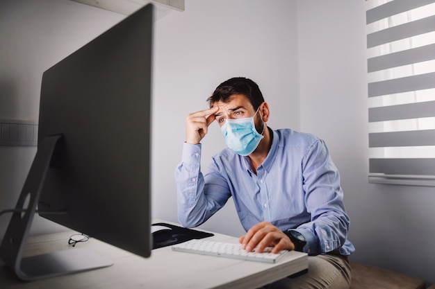 Nervous employee with mask sitting in his office and reading important e- mail during covid outbreak