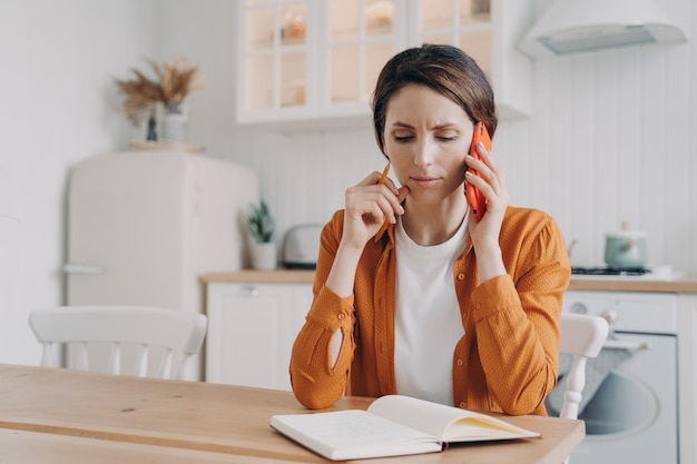 Nervous frowning woman making phone call customer service sitting at table with notebook in kitchen