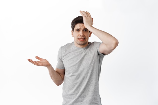 Nervous man look confused and worried, slap forehead and shrugging, forgot remember, made mistake, standing distressed against white background