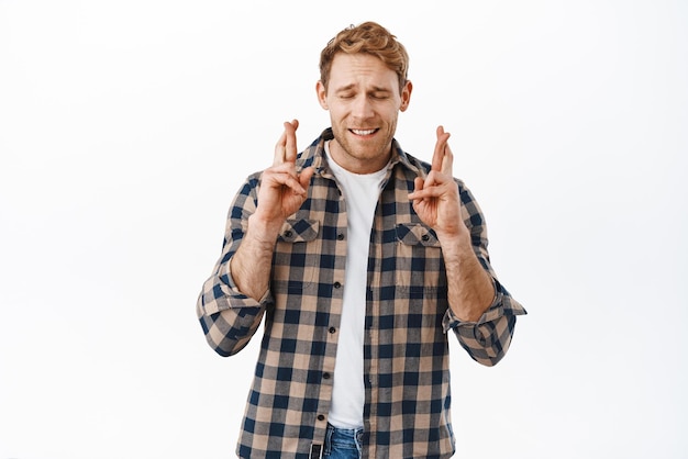 Nervous redhead guy begging god cross fingers for good luck making wish or pleading waiting for news anticipating positive results standing hopeful against white background