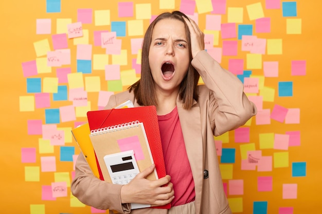 Nervous worried woman holding papers calculating bills standing\
with hand on head forgot important information posing against\
yellow wall with colorful sticky post notes