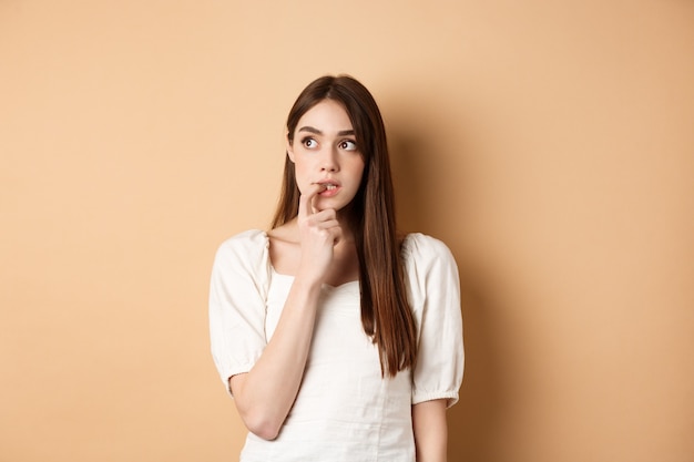 Ragazza giovane nervosa che morde l'unghia e che osserva da parte eccitata, facendo una scelta, pensando a qualcosa, in piedi su sfondo beige.