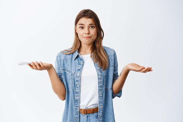 Foto nervosa giovane donna che scrolla le spalle con le mani allargate lateralmente, tenendo il telefono e accigliata turbata, avendo problemi sullo smartphone, in piedi su bianco