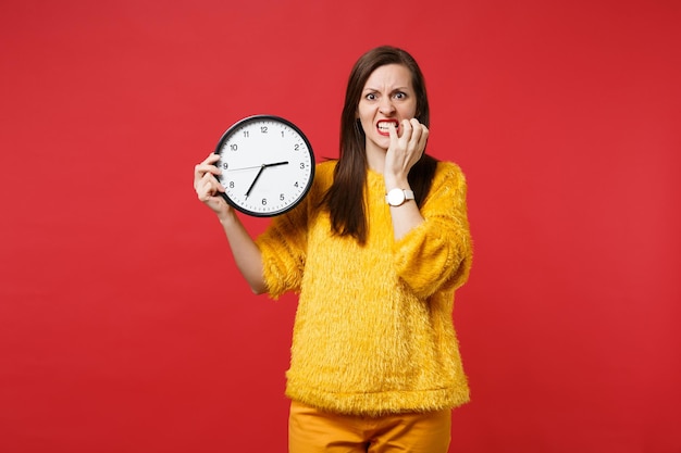 Foto giovane donna nervosa in maglione di pelliccia gialla che rosicchia unghie, tenendo l'orologio rotondo isolato su sfondo rosso in studio. il tempo sta finendo. persone sincere emozioni, concetto di stile di vita. mock up copia spazio.