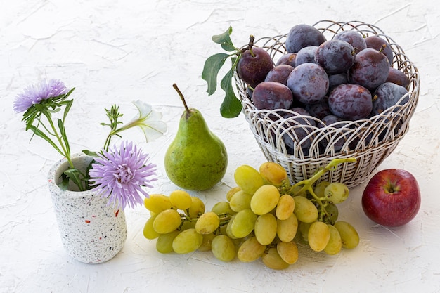 Net rijpe pruimen geplukt in rieten mand, een appel, een peer en een tros rijpe witte druiven op het witte gestructureerde oppervlak