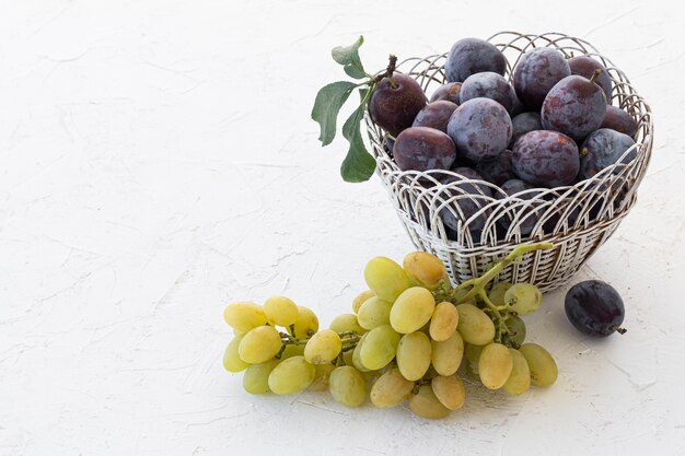 Net rijpe pruimen geplukt in rieten mand en een tros rijpe witte druiven op de witte gestructureerde achtergrond. Net fruit geoogst. Topv-weergave.