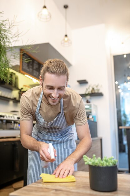 Netheid en netheid. Jonge volwassen vrolijke man in schort afvegen tafel in café met ontsmettingsmiddel