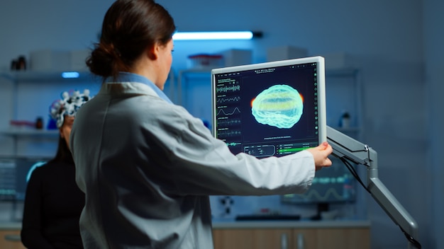 Neurologist doctor reading informations of patient looking at monitor analysing brain scan discussing with patient wearing eeg headset. Scientist taking notes examining nervous system of woman in lab