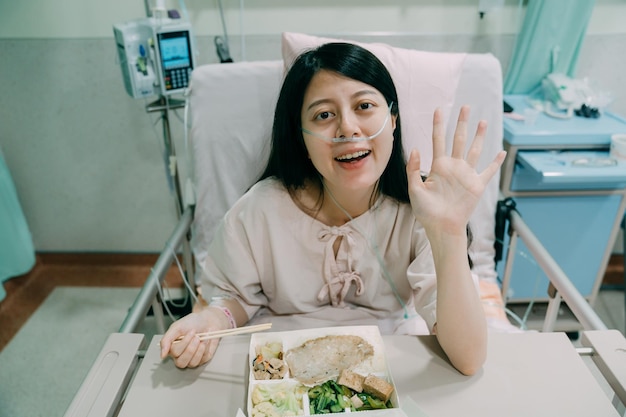 Neuscanule zuurstof therapie concept. jonge aziatische chinese vrouw met borstkanker glimlachend camera kijken met charmant gezicht. vriendelijke dame die met de hand zwaait tijdens het eten van een lunchpakket met stokjes