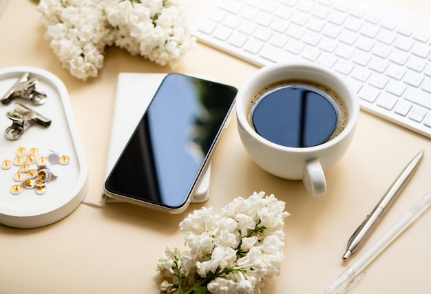 ニュートラルカラーのテーブルトップ、キーボードの白いライラックの花と黒いスマートフォン