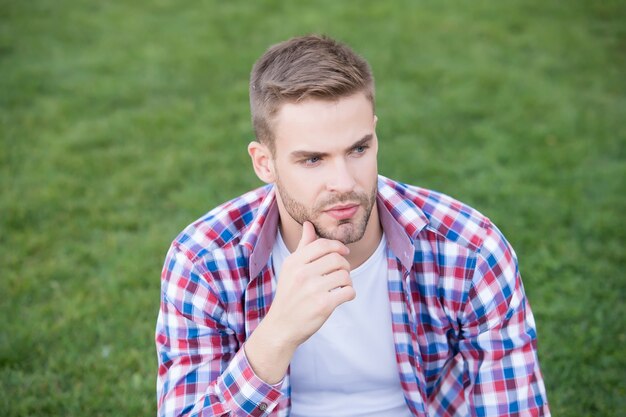 毎日のスキンケアを始めるには若すぎることはありません。若い男は緑の芝生の上に座っています。無精ひげを生やした顔の肌を持つ独身。スキンケア化粧品。適切なスキンケアルーチン。肌の手入れ。メンズスキンケア。