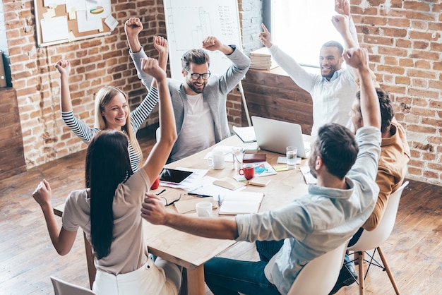 New achievement! Top view of group of six cheerful young people keeping arms outstretched and smile while sitting at the table in office