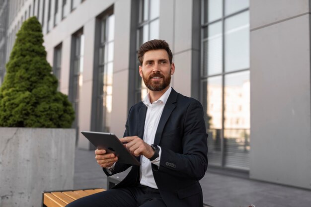 New app ad and offer smiling young european man boss with beard\
in suit use tablet sits on bench