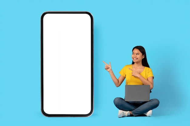 New app for work and study Happy young chinese lady sit with laptop and pointing fingers at smartphone