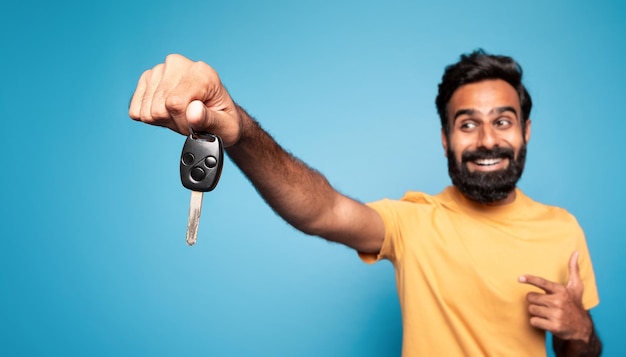 New car happy indian man showing auto key and pointing at it in excitement posing on blue background