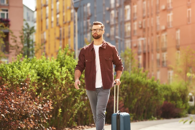 New destination handsome and young bearded man in casual wear and eyeglasses pulling his luggage