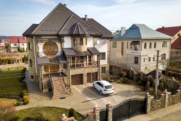 New expensive white car parked in paved well-kept yard with green lawn, decorative bushes and brick fence of big two story house. Luxury and prosperity concept.