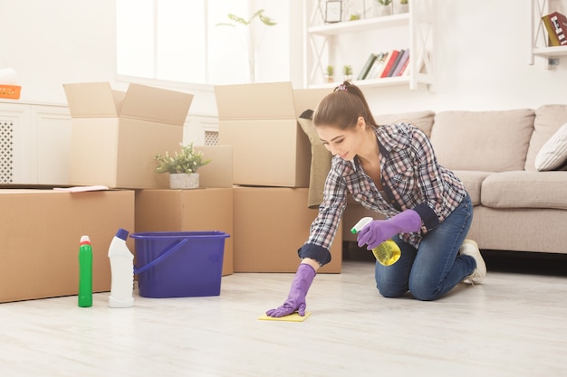 New home cleaning. Young woman tiding up after moving to new appartment. Girl with various detergents, rags and mops in living room full of carton boxes. Household and spring-cleanig concept