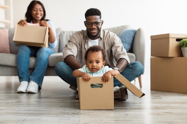 New Home, People And Real Estate Concept. Excited African American family having fun and celebrating moving day, cheerful father riding his little child in cardboard box container in living room