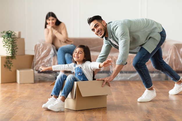 New Home, People And Real Estate Concept. Excited loving family having fun and celebrating moving day, cheerful father riding his little daughter in cardboard box container in living room