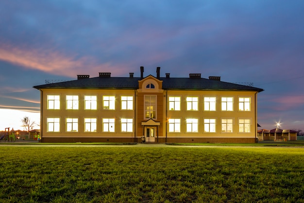 New modern two-storied kindergarten preschool building with brightly lit windows on green grassy lawn and evening blue sky copy space background. Architecture and development concept.