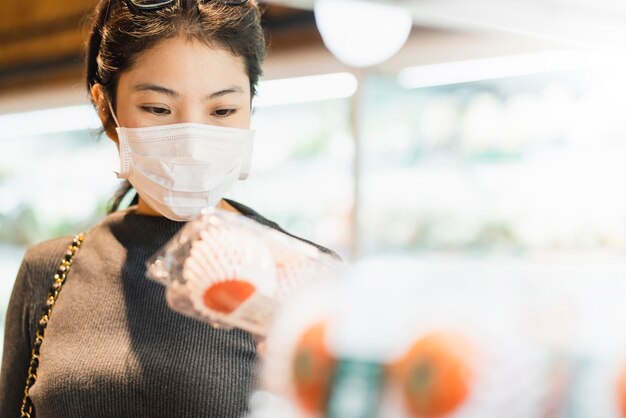 New normal after covid epidemic young smart asian female shopping new lifestyle in supermarket with face shild or mask protection hand choose fresh vegetable of fruit new normal lifestyle